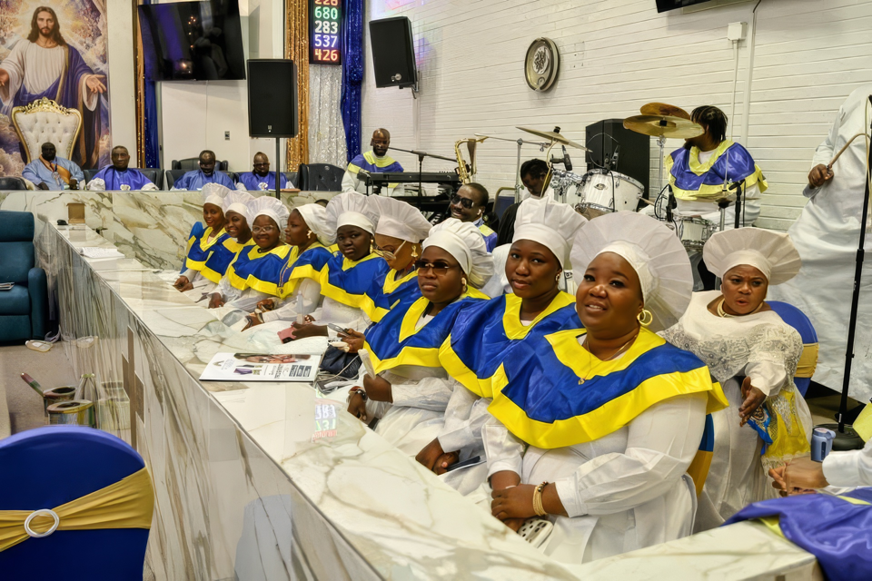 The choir in blue and yellow garments seated beside the musicians, with drums and keyboard behind them.