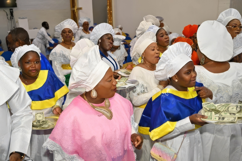 Women in white caps and blue and yellow garments carry offering trays during the thanksgiving procession.