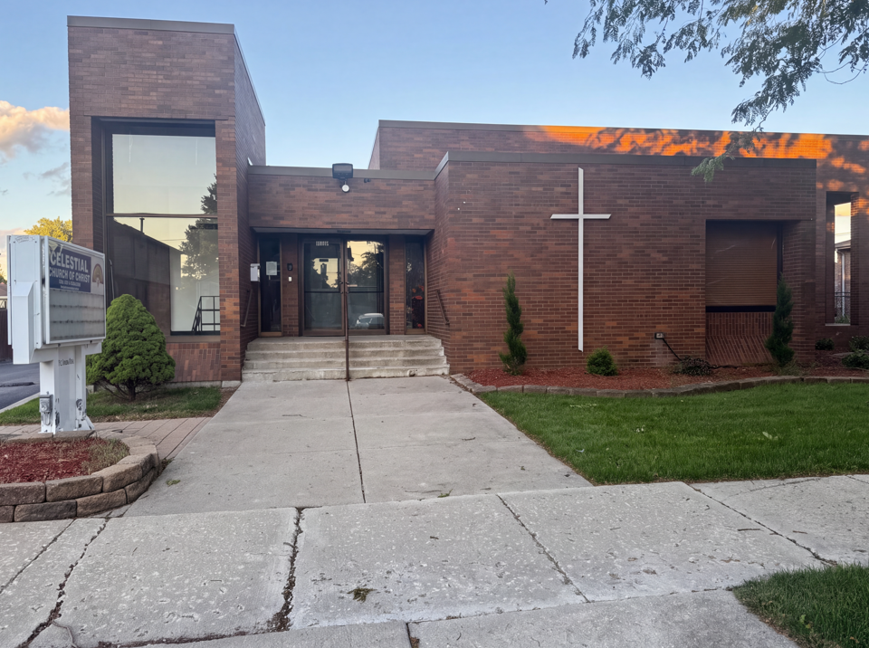 The exterior of SBJ Luli Cathedral, showing its brick facade, white cross, entrance, landscaping, and church sign.