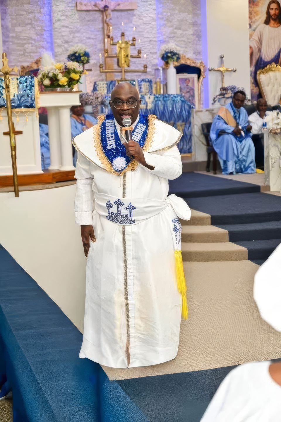 The Shepherd in Charge of SBJ Luli Cathedral, in white garments and a blue sash reading Shepherd in Charge, addressing the congregation with a microphone near the altar steps.